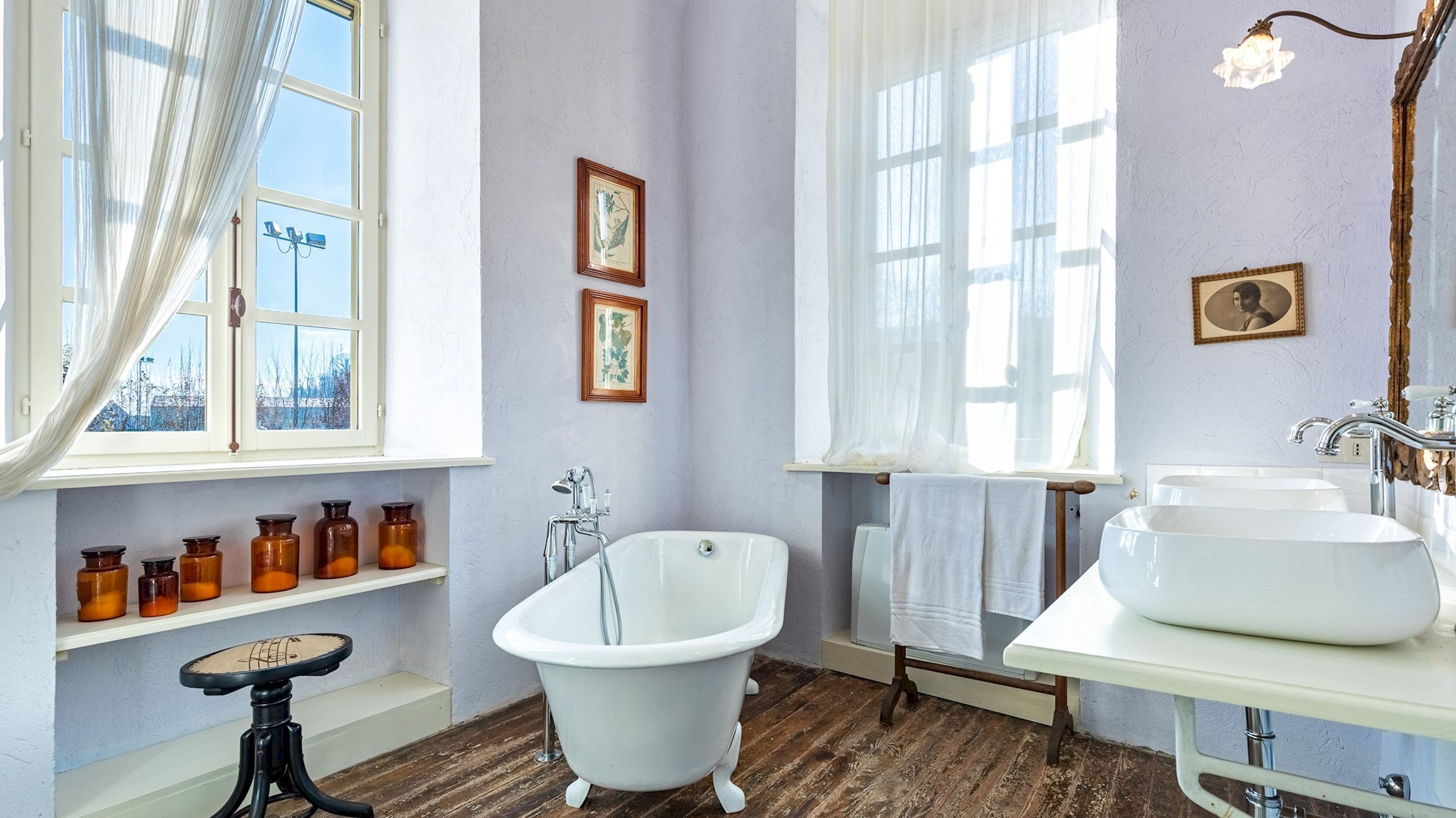 A bathroom with a clawfoot bathtub, a large window with sheer curtains, and a wooden shelf with jars.