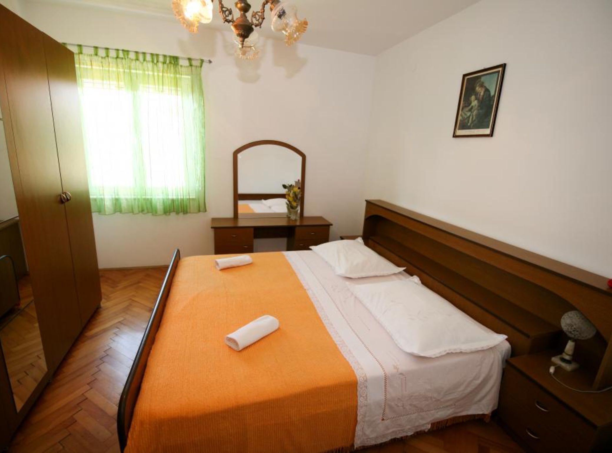 Habitación de hotel con cama de madera, cómoda, espejo, un cuadro en la pared y ventana con cortinas verdes.