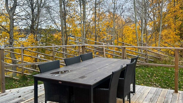 Une terrasse dotée d’une table et de chaises en bois foncé, entourée d’une balustrade en bois et avec, en arrière-plan, le feuillage automnal.