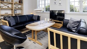 A modern living room with a black leather sofa, a wooden coffee table, and a television on a wooden stand. There are shelves with decorative items and a window with a view of greenery. - 4 Star Holiday Home in Saeby (Sæby)