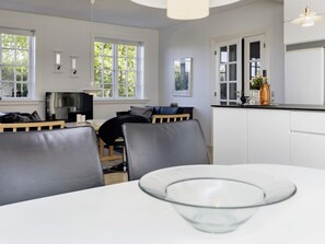 Una cocina moderna con armarios blancos, un comedor con un bol de vidrio sobre la mesa, y una zona de estar con televisor y lámpara.