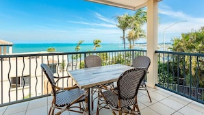 Ein Balkon mit Tisch und Stühlen, der Blick auf einen Strand mit Palmen und einem strahlend blauen Himmel bietet.