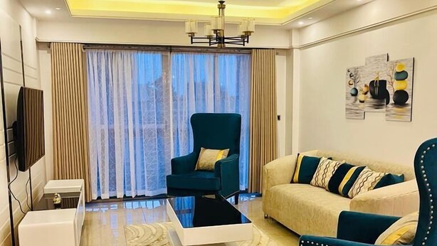 A modern living room with a sofa, two armchairs, a coffee table, a TV, and a large window with curtains. - Vesta Garden Apartments Kilimani (Nairobi)