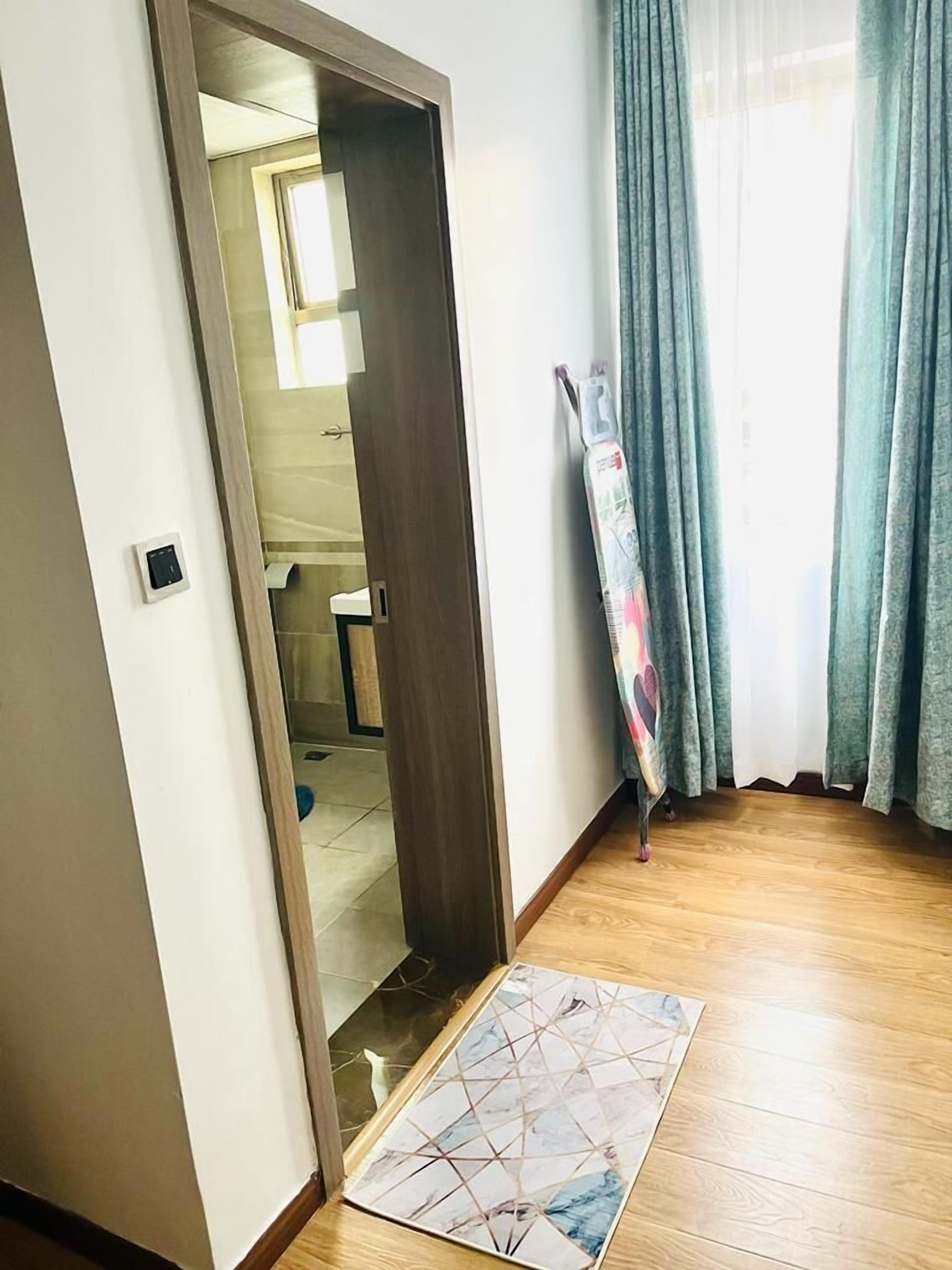 A room with a wooden floor, a door leading to a bathroom, and a window with curtains.