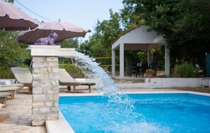 A stone fountain with water spouting into a swimming pool, surrounded by lounge chairs and umbrellas. - Spacious Prgomet Retreat With Private Pool, Fitness Centre, and Garden (Prgomet)