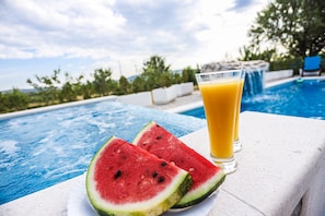 A swimming pool with a glass of orange juice and two slices of watermelon on a white surface. - Secluded Holiday Home With Pool and Hot Tub (Lecevica)