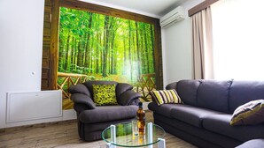 A living room with a large forest mural, two sofas, a glass coffee table, and a radiator.