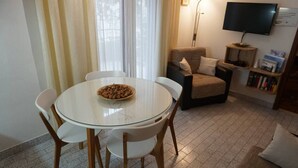 A dining area with a round glass table and white chairs, a bowl of nuts on the table, a sofa, a TV on a stand, and a bookshelf with books. - Modern Trogir Apartment With Private Patio, Bbq, and Close to Beach (Trogir)