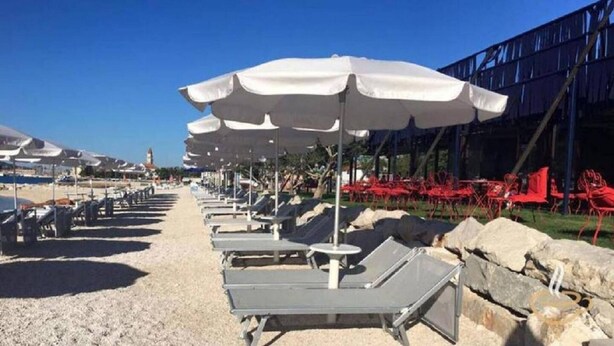 A beachside area with rows of sun loungers and white umbrellas, a rocky shoreline, and a building in the background. - Modern Trogir Apartment With Private Patio, Bbq, and Close to Beach (Trogir)