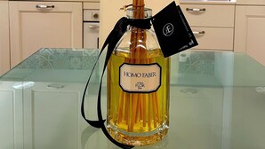 A glass bottle with a label reading "HOMO FABER" and a tag, placed on a glass table in a kitchen with patterned tiles and white cabinets.