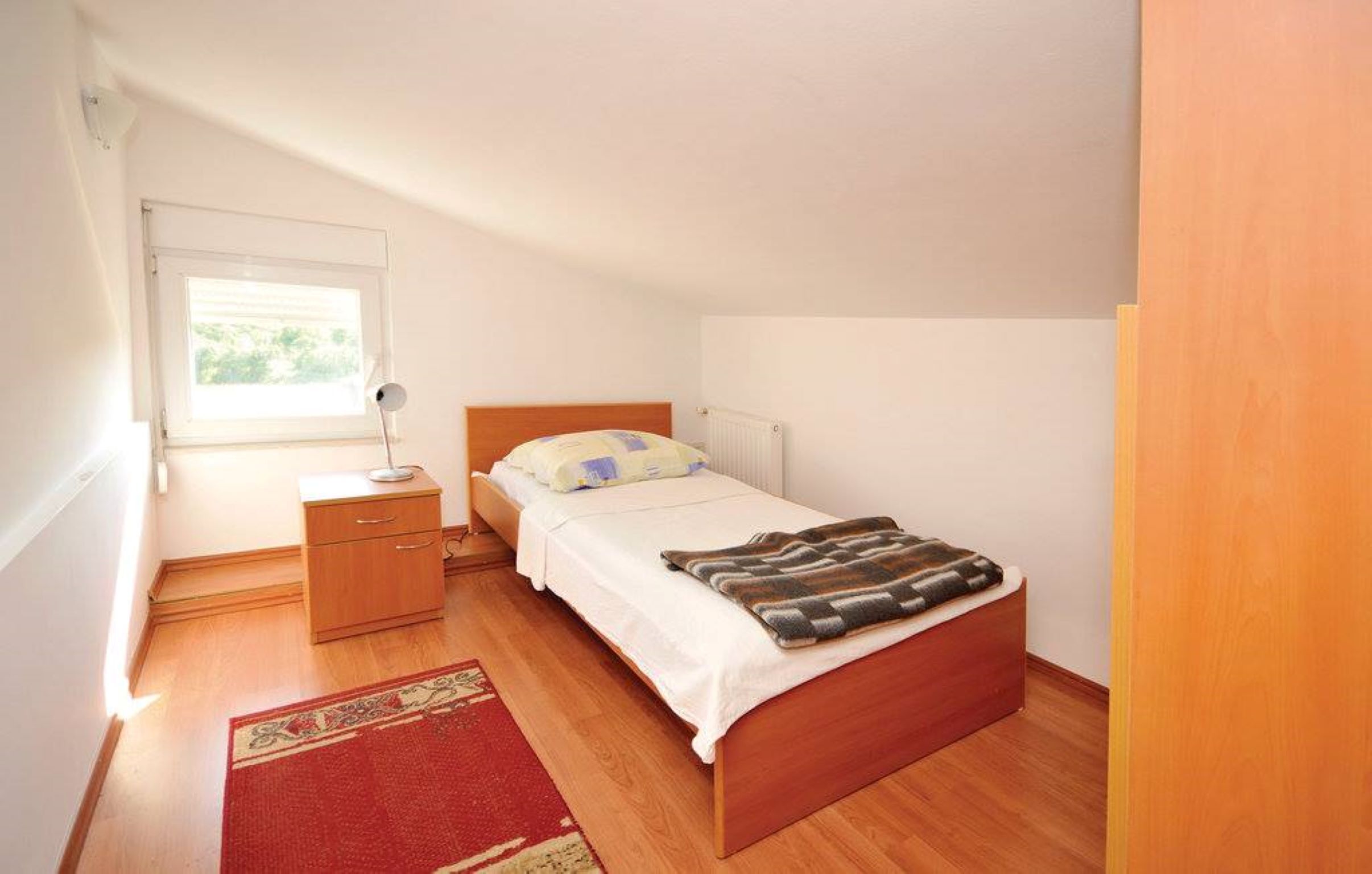 A bedroom with a single bed, a bedside table, a nightstand, a window, and a red rug.