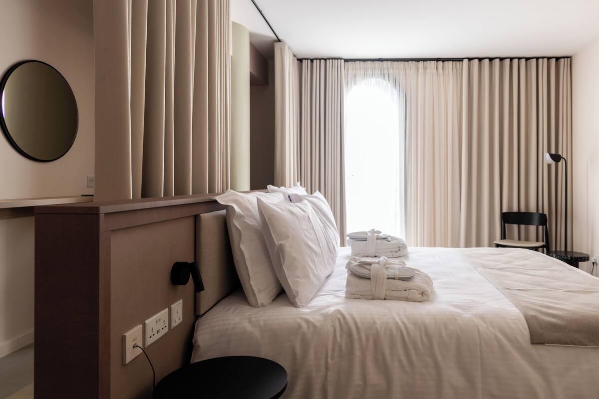 Ein Hotelzimmer mit einem ordentlich bezogenen Bett, einem runden Spiegel, einem Nachttisch und einer Lampe.