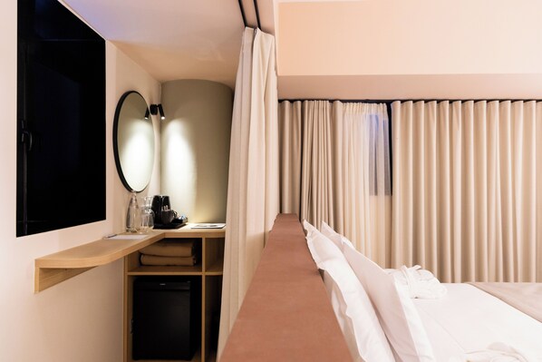 Quarto de hotel com cama, escrivaninha com espelho, prateleira com artigos de higiene e janela com cortinas.