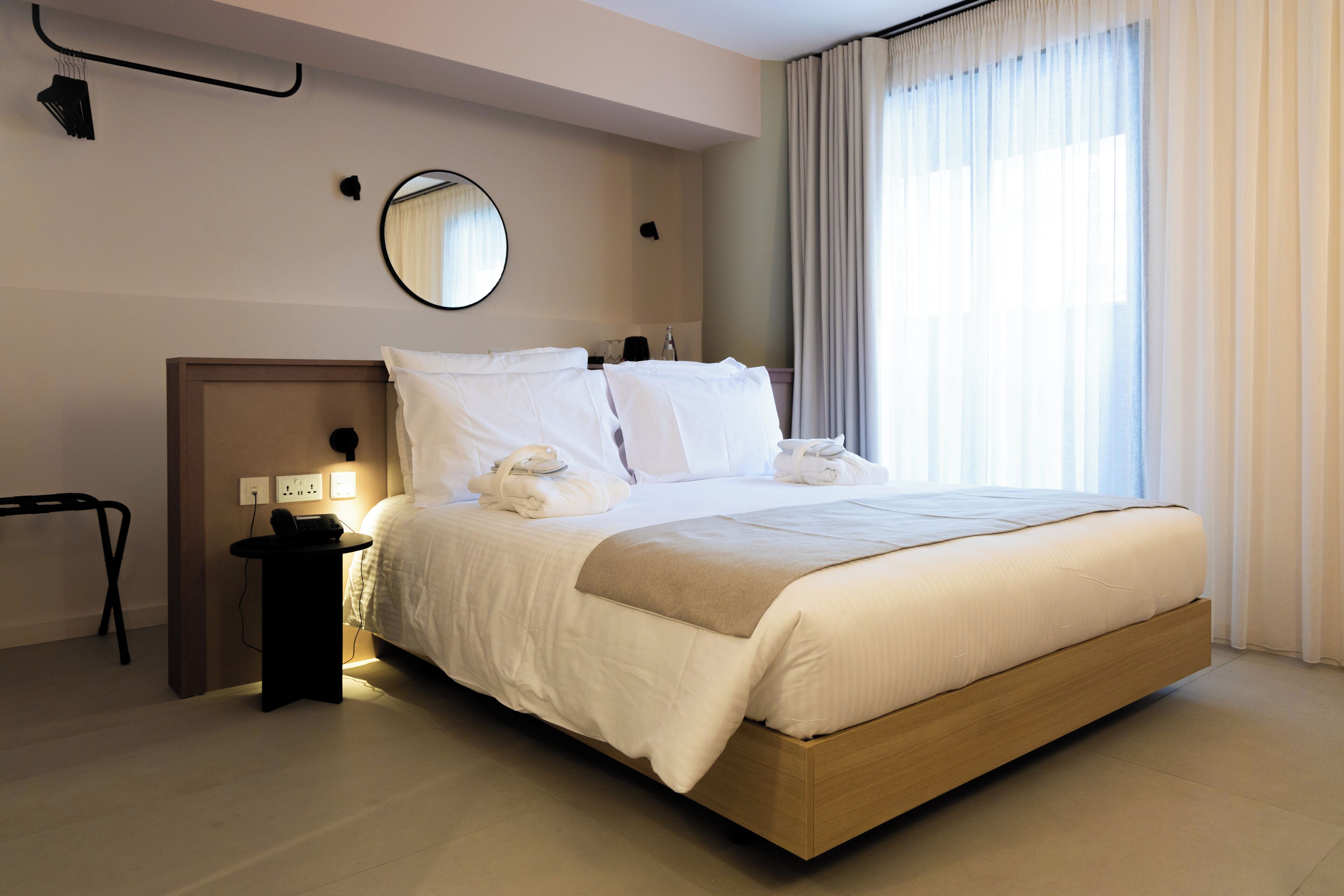 A modern hotel room with a large bed, bedside tables, a mirror, and a view of the outdoors through sheer curtains.