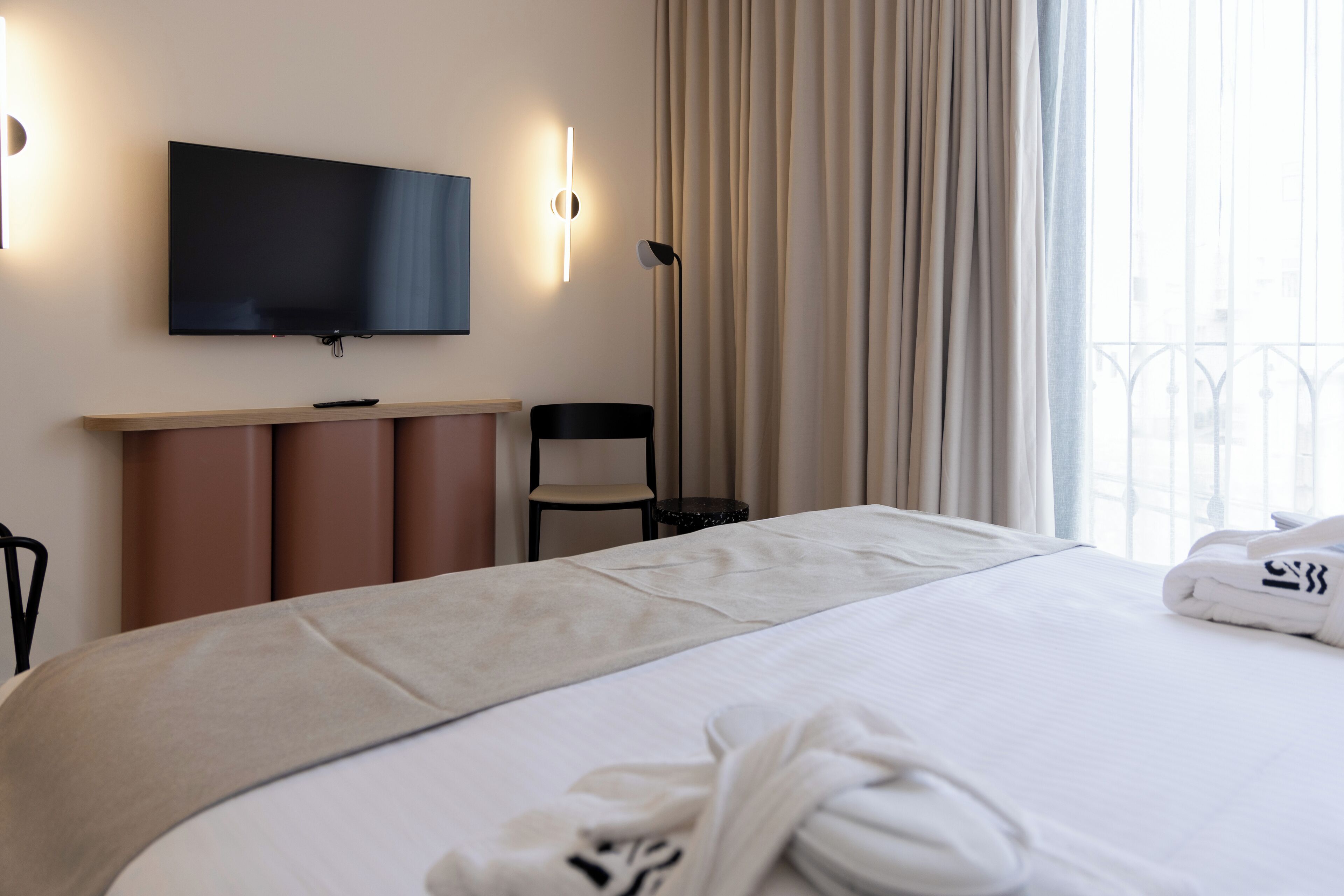 A hotel room with a large bed, a flat-screen TV mounted on the wall, a desk with a chair, and a window with curtains.