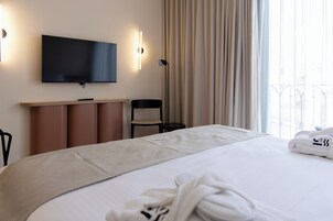 A hotel room with a large bed, a flat-screen TV mounted on the wall, a desk with a chair, and a window with curtains.
