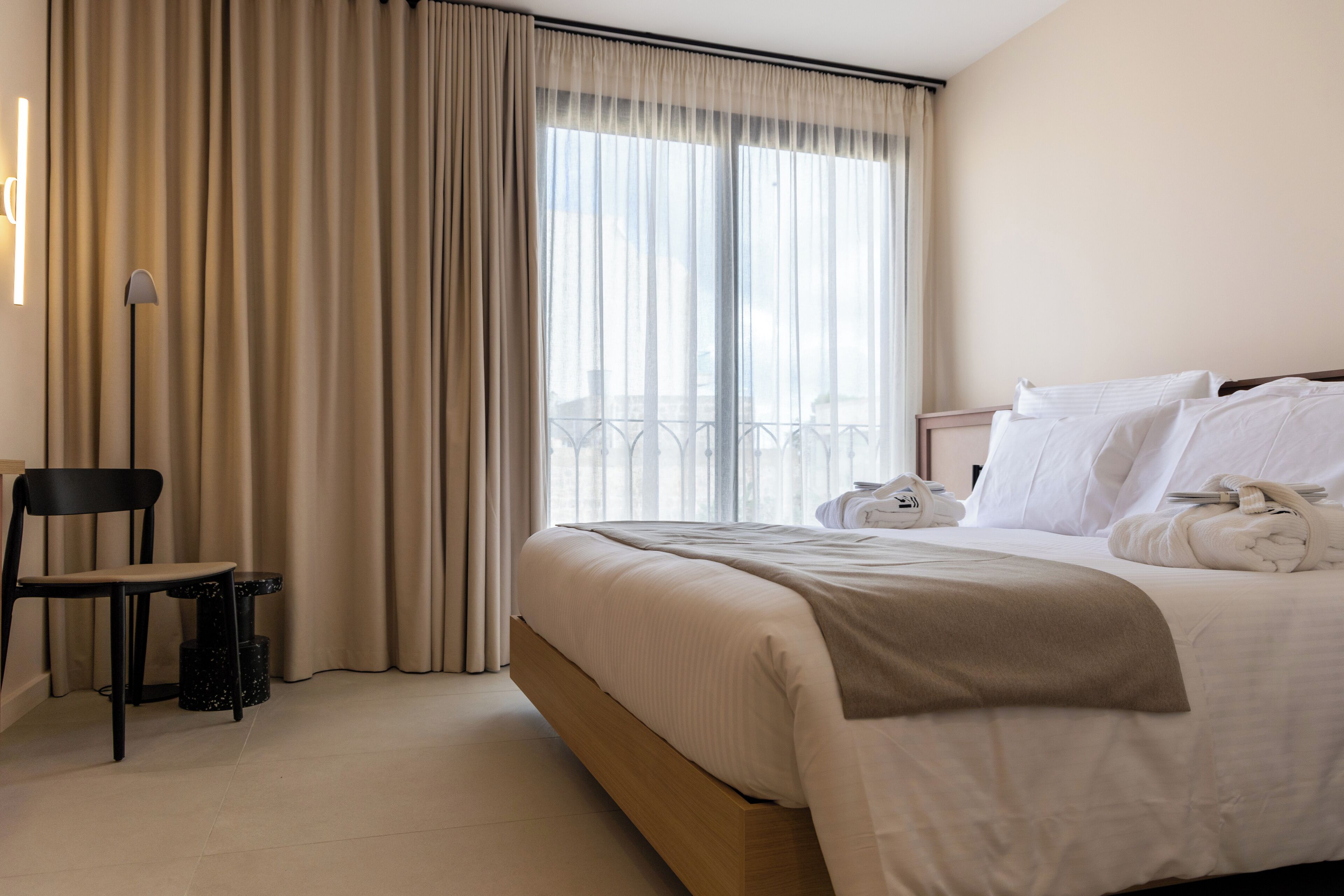 Ein Hotelzimmer mit Bett, Sessel, Lampe und Blick ins Freie durch große Fenster.