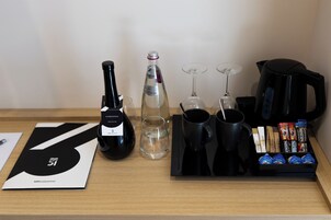 Habitación de hotel con una mesa de madera que muestra una botella de vino, copas, una cafetera y un bandeja con cápsulas de café.