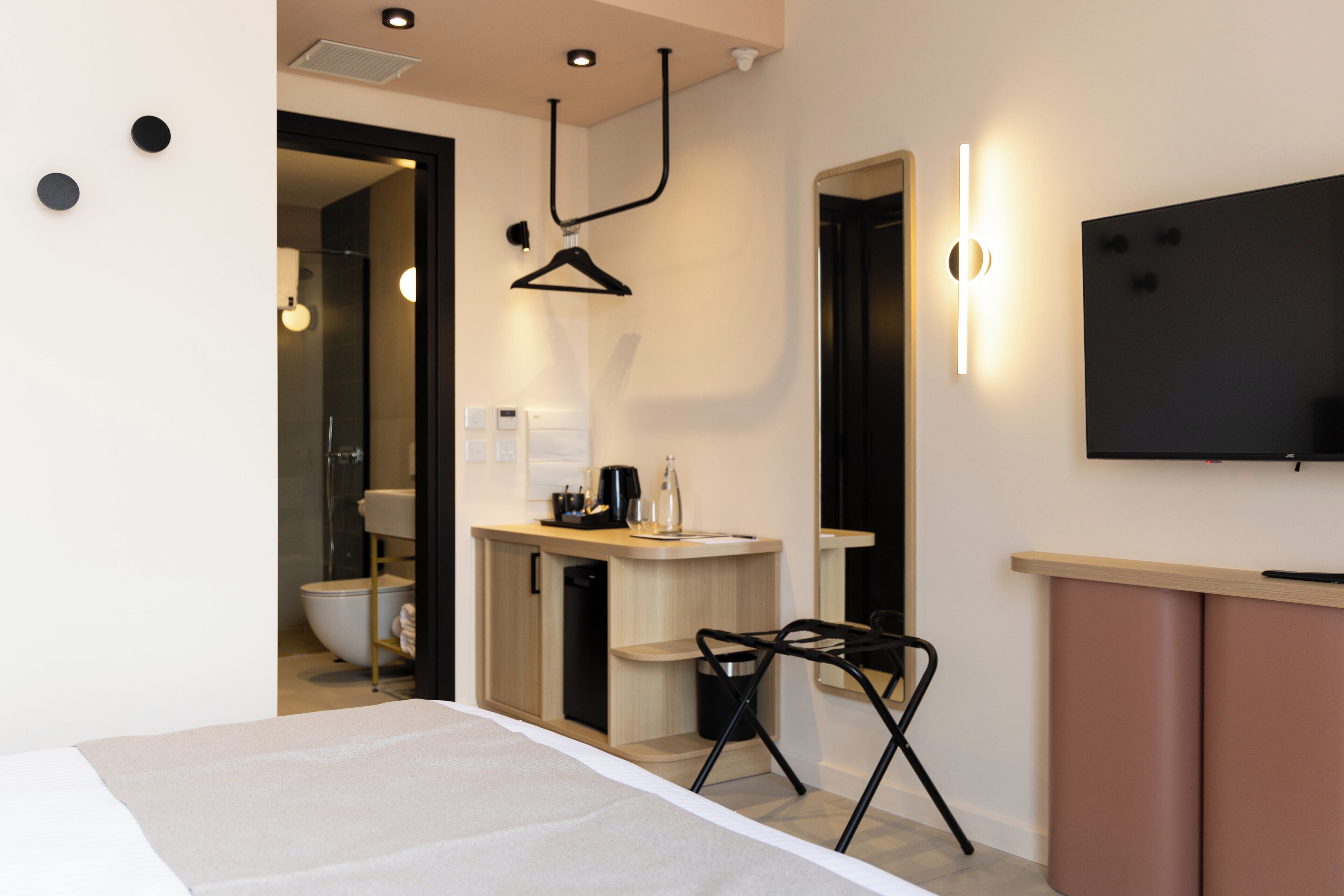 Une chambre d’hôtel moderne, équipée d’un lit, d’un petit bureau avec une chaise, d’une télévision et d’une salle de bain visible à travers une porte ouverte.