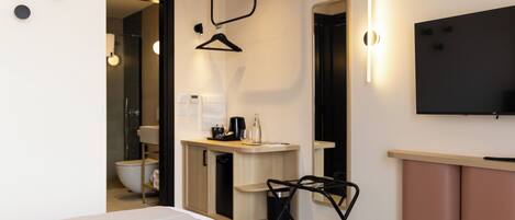 Une chambre d’hôtel moderne, équipée d’un lit, d’un petit bureau avec une chaise, d’une télévision et d’une salle de bain visible à travers une porte ouverte.