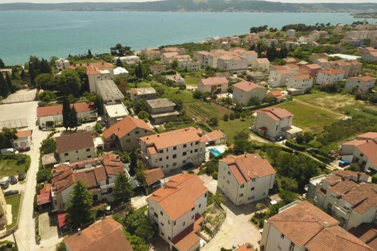 A residential area with multiple two-story houses, some with red-tiled roofs, surrounded by greenery and a body of water in the background.