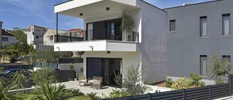 A modern two-story building with a balcony, a covered outdoor seating area, and a fenced garden.