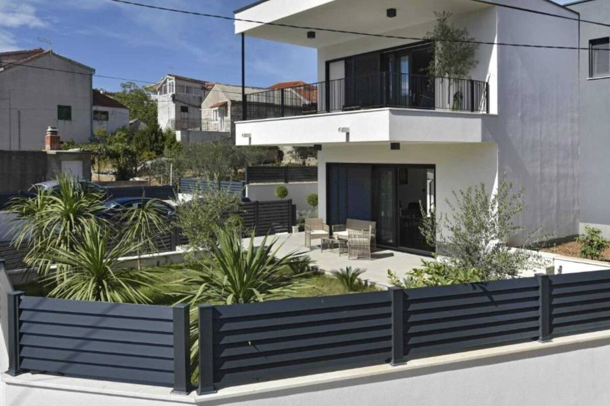 A modern two-story house with a balcony, a patio with outdoor furniture, and a fenced garden area.