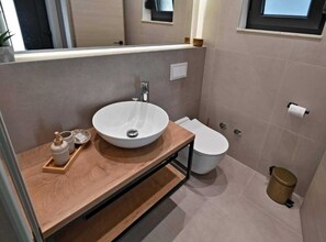 A modern bathroom with a wooden vanity, a white sink, a toilet, and a trash can. - Luxury Slatine Apartments With Terrace and Garden (Split)