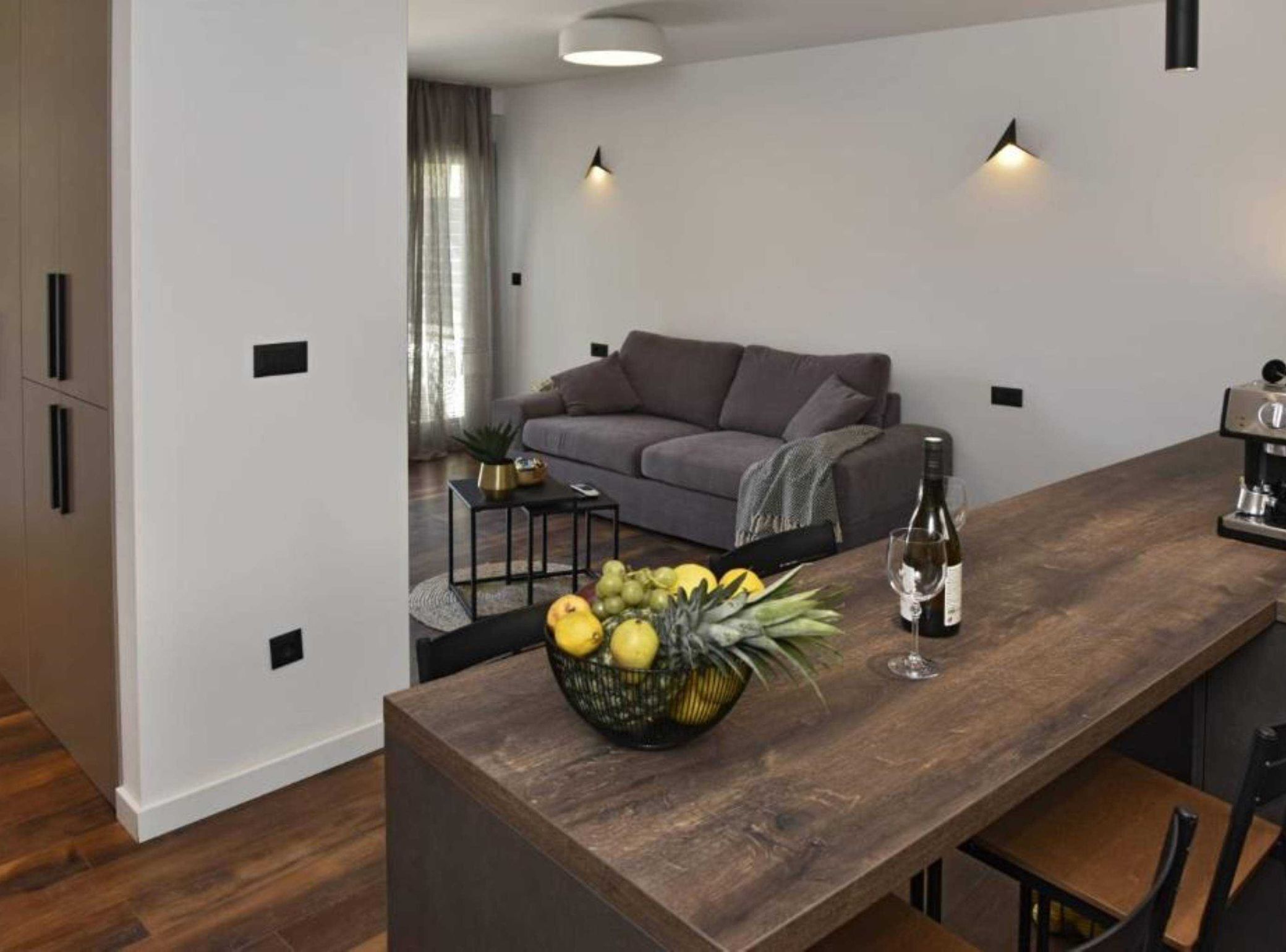 A modern living room with a grey sofa, a wooden dining table with a fruit bowl, and a coffee machine.