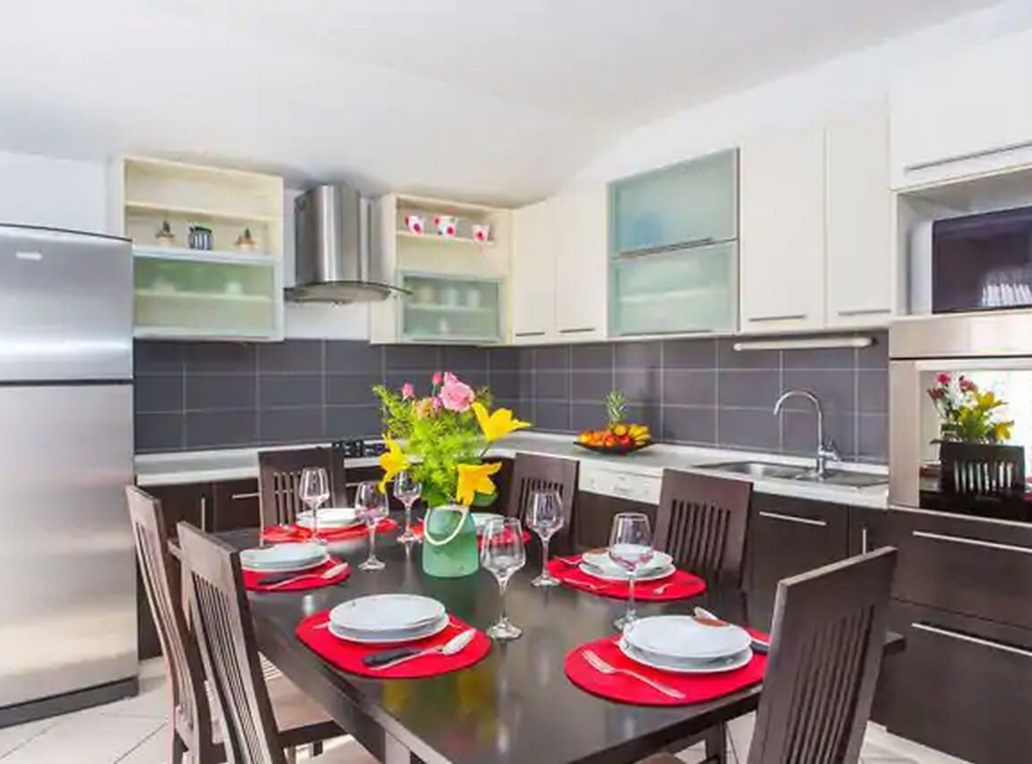 A modern kitchen with a dining table set for four, equipped with red placemats and glassware.
