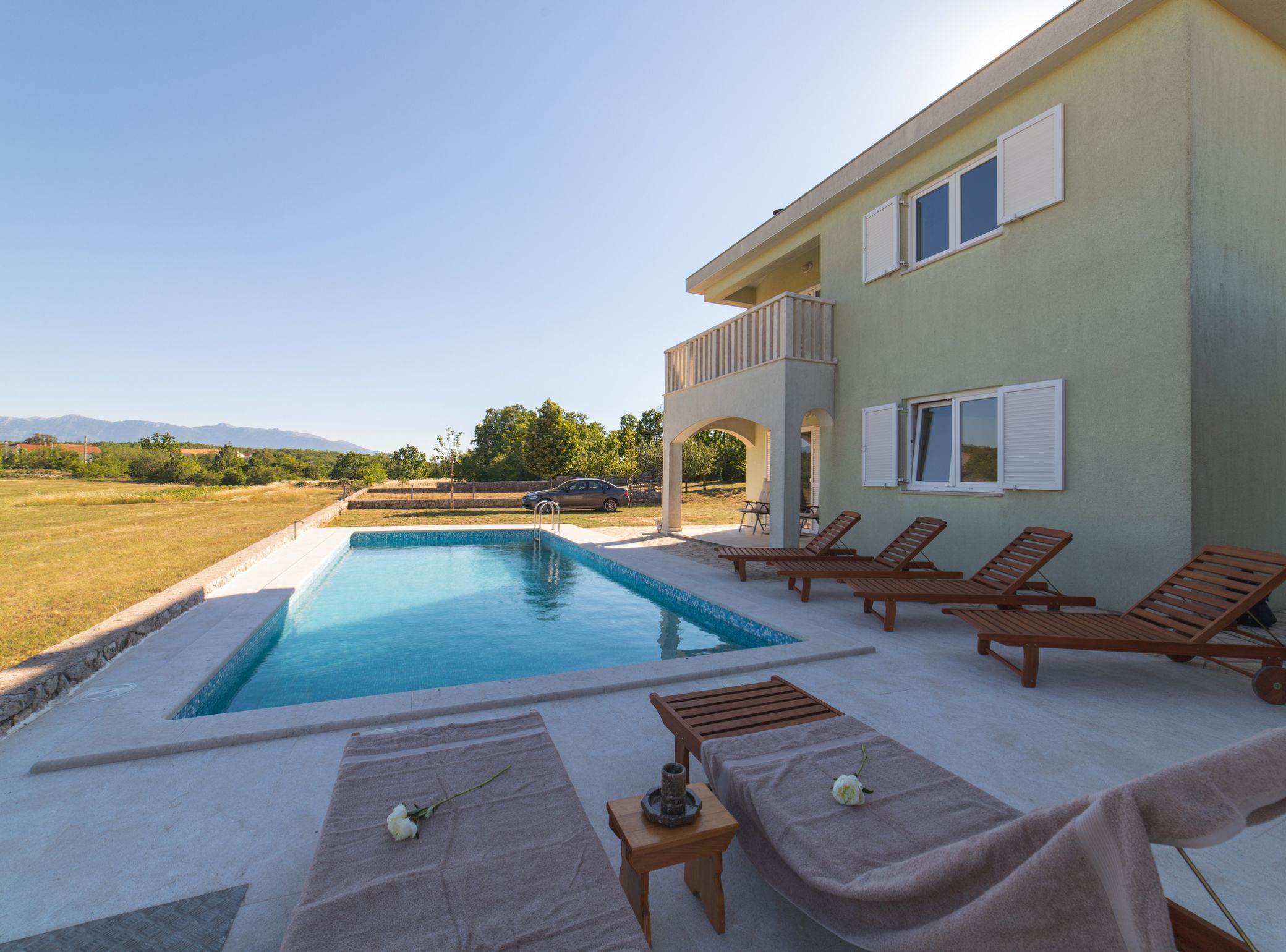 Une piscine avec des chaises longues et une table, une voiture garée à proximité, et un immeuble doté d’un balcon.