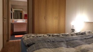 A bedroom with a bed, a wooden wardrobe, a bathroom with a mirror, and a bedside lamp. - Lovely Apartment With Mountain Views (Veysonnaz)