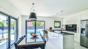 A modern kitchen with a central island, a dining area with a table and chairs, and a large window providing a view of the outdoors. - Modern Kastel Villa With Private Pool, Gym, and Large Outdoor Space (Kastela)