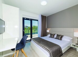 Ein modernes Hotelzimmer mit einem großen Bett, einem Schreibtisch und einem Stuhl. Ein Balkon bietet Blick auf Grünflächen und Gebäude.