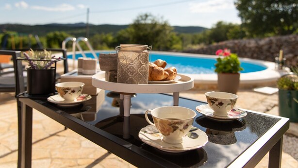 A table set with tea cups, a teapot, and pastries by a poolside with a scenic view. - Serene Retreat Holiday Home With Pool (Klis)