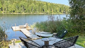 A lakeside deck with a table and two chairs, a dock with a boat, and a forested background. - 4 Star Holiday Home in Ljusfallshammar (Ljusfallshammar)