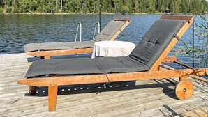 A wooden deck with two sun loungers, one with a table, and a forested shoreline in the background. - 4 Star Holiday Home in Ljusfallshammar (Ljusfallshammar)