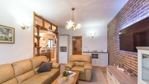 Un salon chaleureux avec un canapé, un fauteuil et une table basse, un coin cuisine avec un mur en briques, et un espace repas avec des chaises et une table.