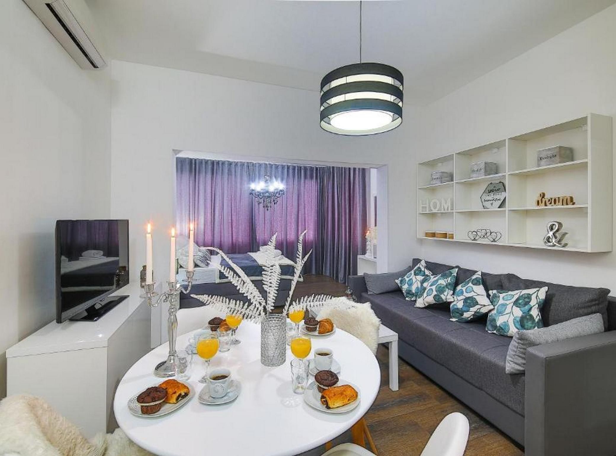 A modern living room with a dining table set for breakfast, a grey sofa, and a white shelving unit with decorative items.