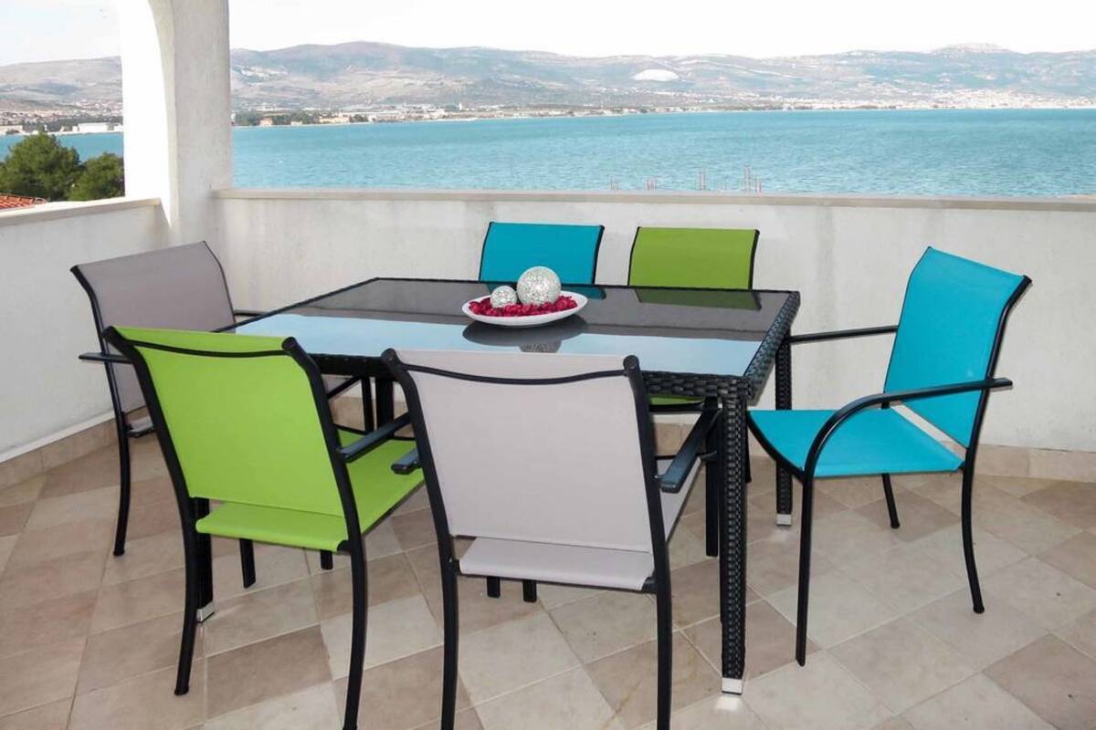 A terrace with a glass table and chairs, a plate with decorative items, and a view of the sea and distant hills.
