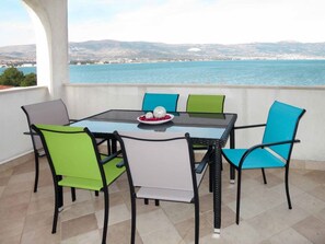 A terrace with a glass table and chairs, a plate with decorative items, and a view of the sea and distant hills.