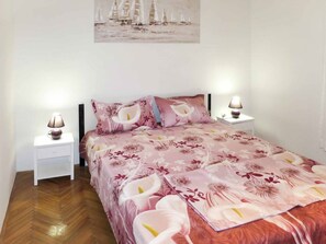 A bedroom with a floral bedspread, two bedside lamps, a nightstand, and a painting of sailboats on the wall.