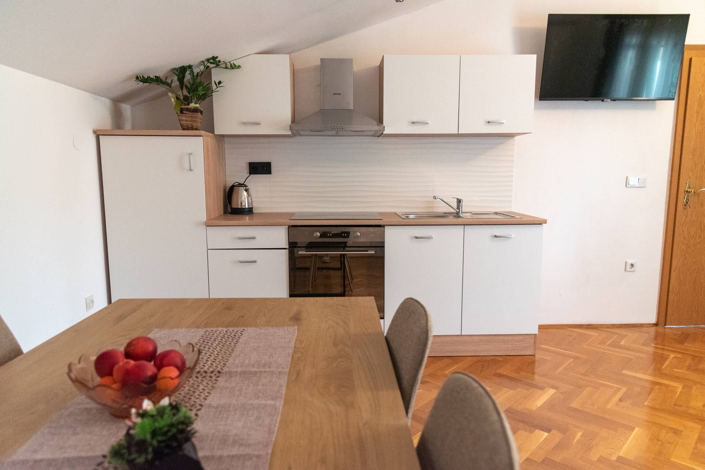 Eine moderne Küche mit weißen Schränken, einer Edelstahl-Dunstabzugshaube und einem eingebauten Backofen. Ein Esstisch mit Obstschale und Blumentopf steht daneben.
