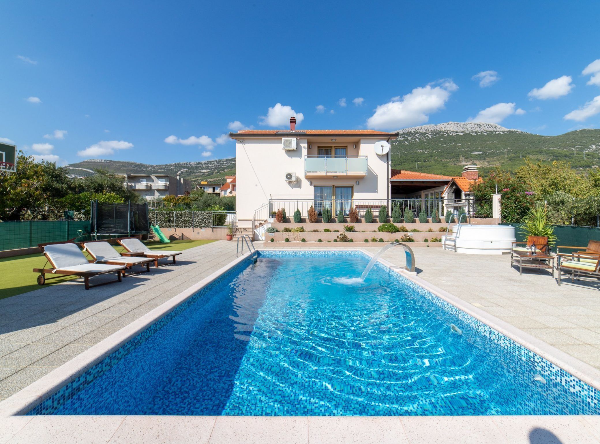 A swimming pool with lounge chairs, a building with balconies, and a mountainous backdrop.