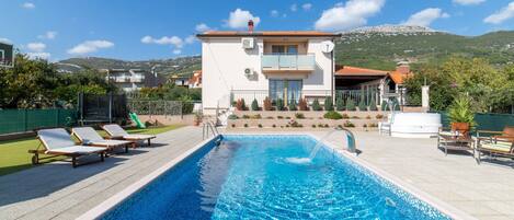 A swimming pool with lounge chairs, a building with balconies, and a mountainous backdrop.