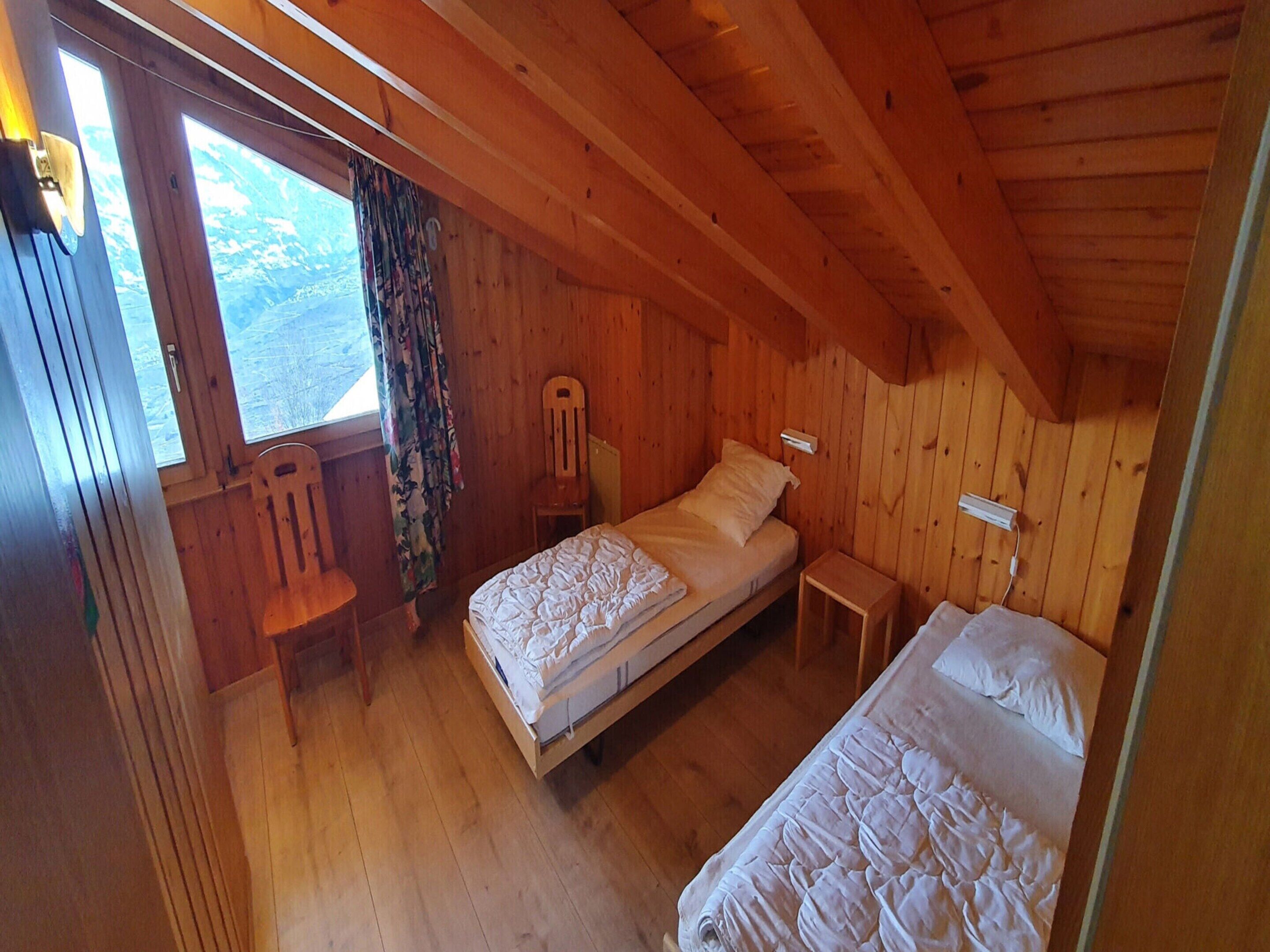 A wooden cabin bedroom with two beds, a chair, a nightstand, and a window with curtains.