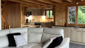 A modern cabin interior with a cozy living area featuring a white leather sofa with black and white cushions, a wooden ceiling, and a kitchen with wooden cabinets and a window overlooking greenery. - Mont Bijou Mountain & Quiet Chalet 10 Pers (Veysonnaz)