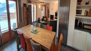 A cozy, well-lit room with a wooden dining table, comfortable chairs, a built-in kitchen with a refrigerator, microwave, and coffee maker, and a large window offering a view of the outdoors. - Mont Bijou Mountain & Quiet Chalet 10 Pers (Veysonnaz)