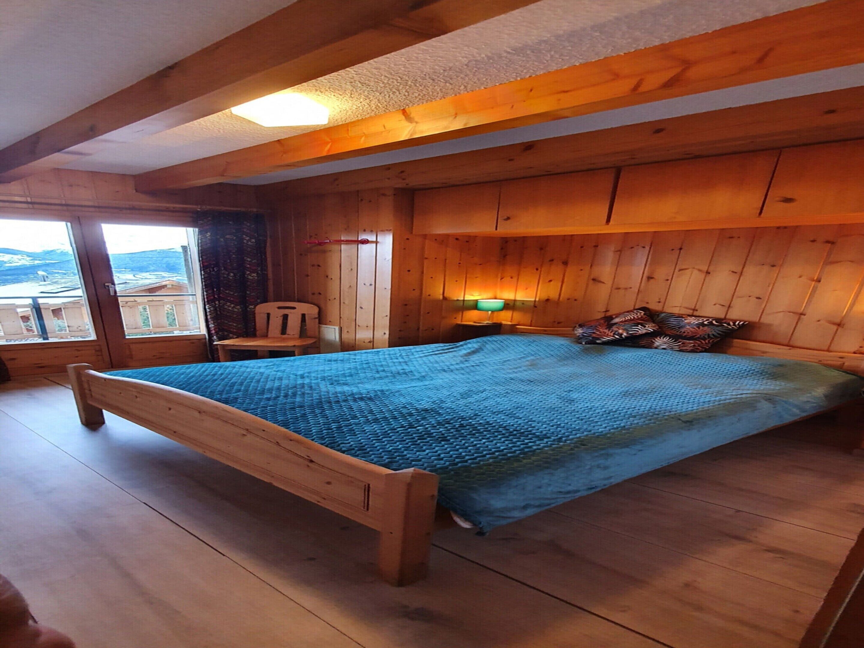 A wooden bedroom with a bed, a chair, a nightstand, and a window with a view.