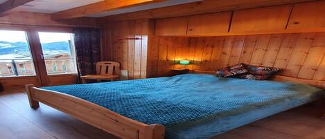 A wooden bedroom with a bed, a chair, a nightstand, and a window with a view.